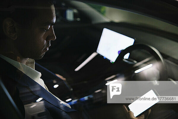 Businessman using smart phone sitting in electric car seen through window