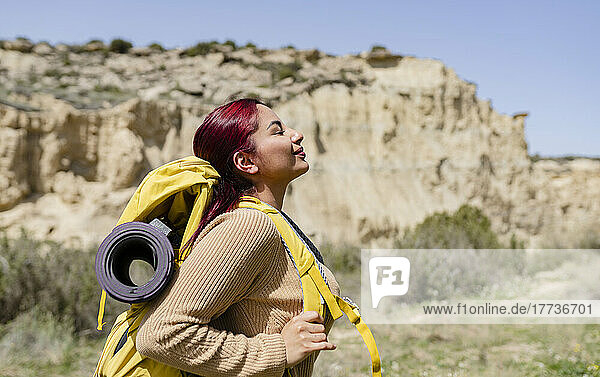 Lächelnde Frau mit Rucksack steht an einem sonnigen Tag mit geschlossenen Augen