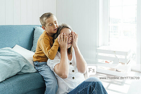 Boy covering eyes of mother sitting by sofa at home
