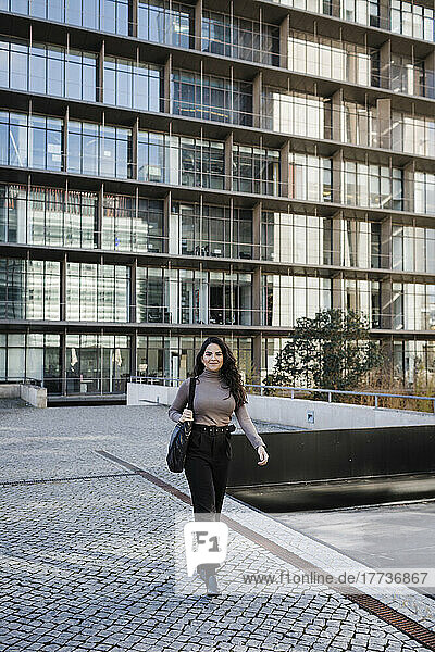 Lächelnde junge Geschäftsfrau  die vor einem modernen Gebäude im Finanzviertel läuft
