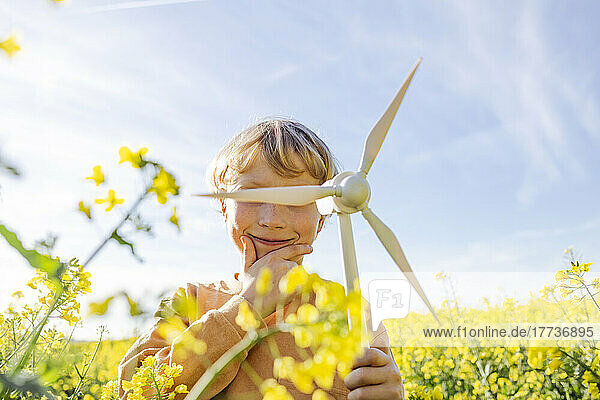 Boy with hand on chin holding wind turbine model at rapeseed field