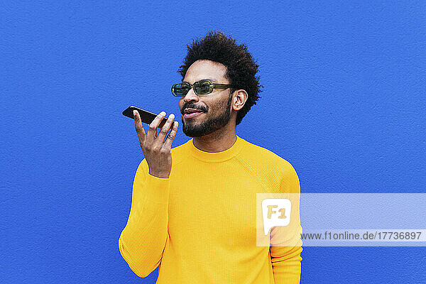 Smiling man talking on mobile phone through speaker in front of blue wall