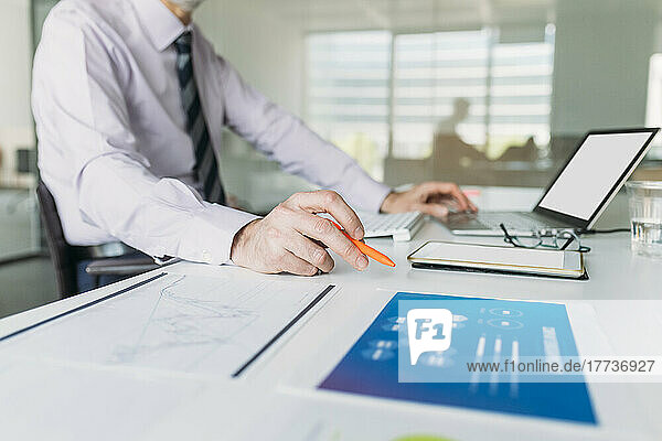 Businessman holding pen sitting with laptop at desk in office