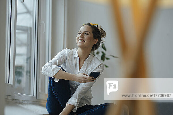 Happy woman holding mobile phone sitting by widow at home