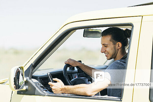 Smiling young man using smart phone sitting in van on sunny day