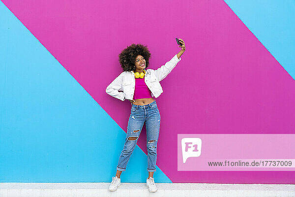 Smiling woman with hand on hip taking selfie through mobile phone in front of pink and blue wall