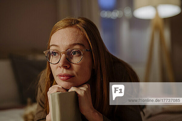 Thoughtful redhead woman wearing eyeglasses lying with book on bed at home