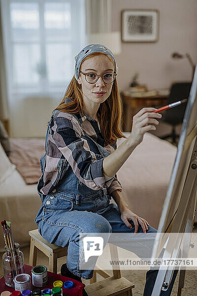 Frau mit Brille sitzt zu Hause auf einem Hocker neben der Leinwand des Künstlers