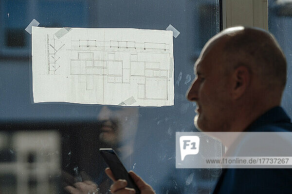 Businessman with mobile phone by blueprint on window in office