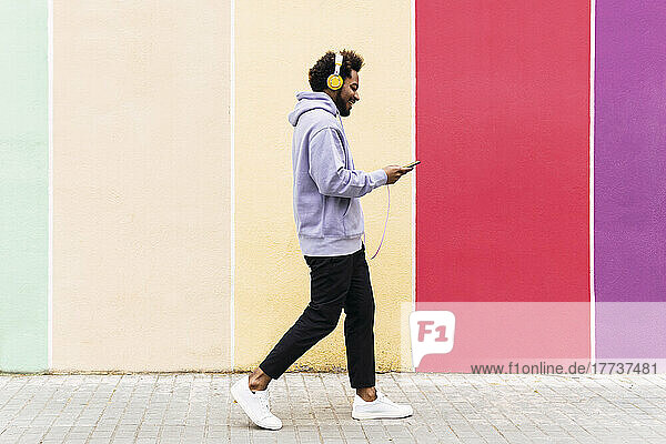 Smiling man using mobile phone and listening music through headphones walking on footpath
