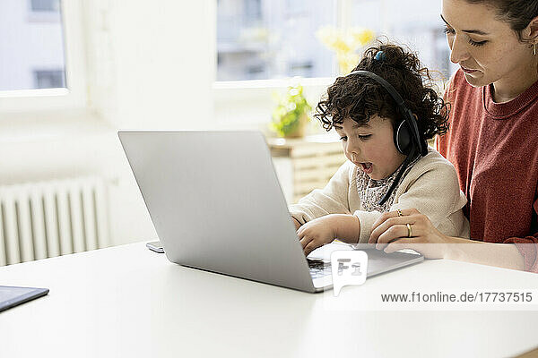 Little girl with headset sitting on lap of mother using laptop
