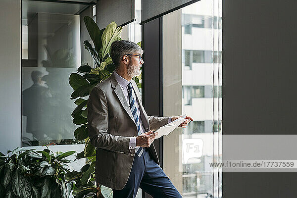 Businessman with tablet PC and documents looking through window in office