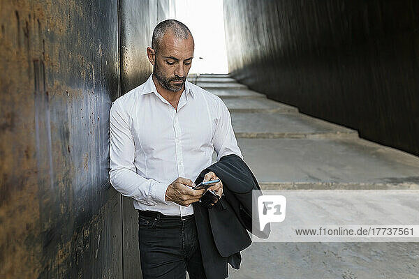 Man using smart phone leaning against wall in alley