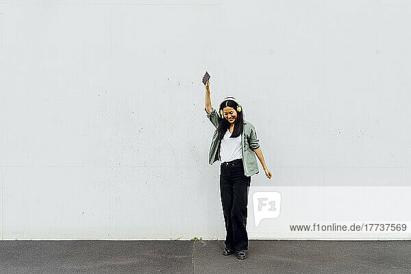 Lächelnde Frau mit erhobener Hand genießt Musik über kabellose Kopfhörer vor einer weißen Wand
