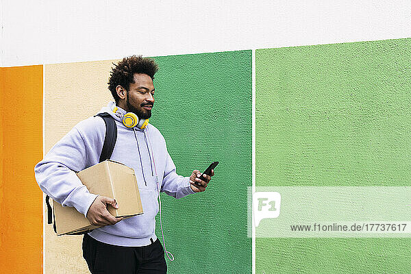 Lieferbote hält Karton mit Smartphone vor bunter Wand