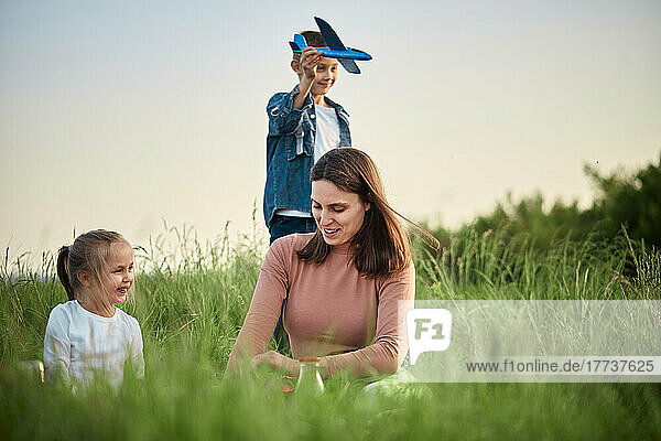 Lächelnde Mutter und Tochter vor einem Jungen  der mit einem Spielzeugflugzeug auf dem Feld spielt