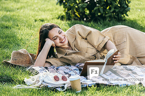 Happy young woman reading book lying on picnic blanket at park