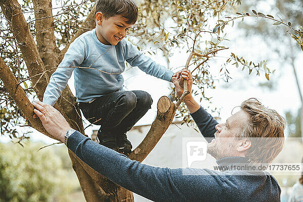 Father holding hands of son crouching on tree