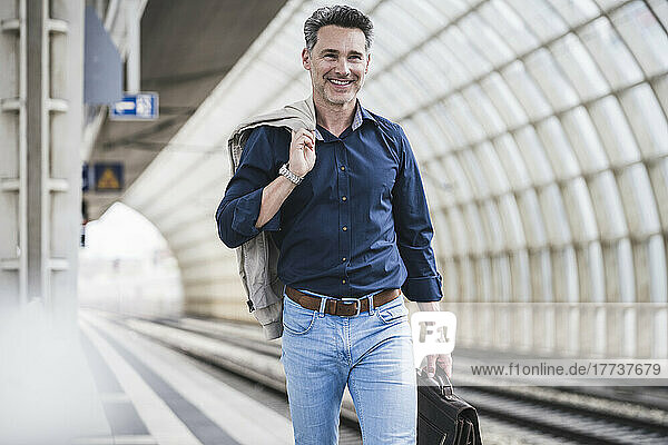 Smiling mature businessman carrying jacket and bag walking at railroad station