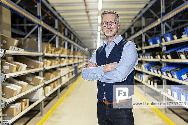 Smiling confident manager with arms crossed standing in aisle at warehouse