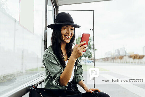 Smiling young woman surfing net through smart phone sitting at bus stop