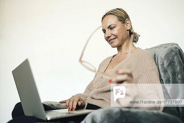 Smiling businesswoman holding eyeglasses using laptop on armchair at home