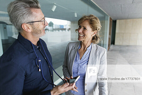 Smiling businessman holding smart phone discussing with businesswoman in front of glass wall