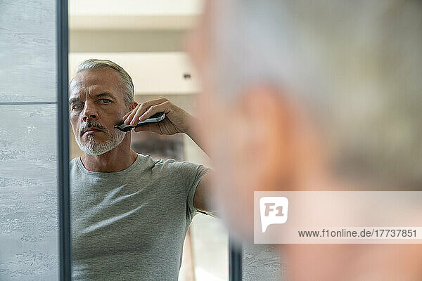 Mann rasiert Bart mit Elektrorasierer und schaut in den Spiegel