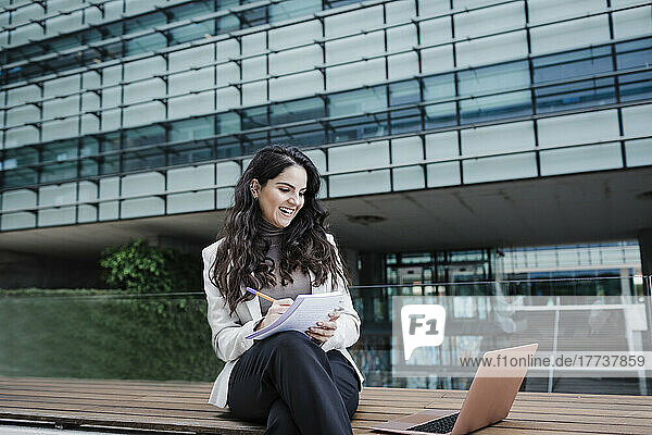 Glückliche junge Geschäftsfrau schreibt in ein Buch und schaut auf den Laptop