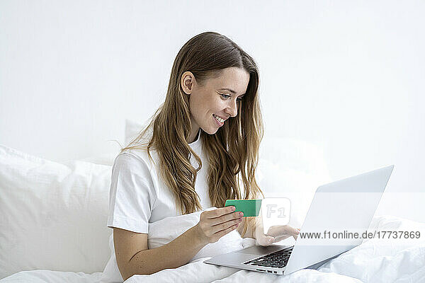 Happy woman holding credit card doing online shopping through laptop in bedroom at home