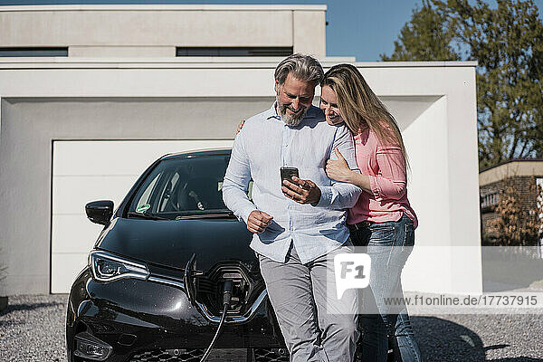 Smiling man sharing mobile phone with woman in front of electric car