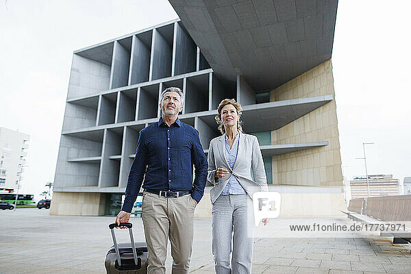 Businessman with luggage walking by businesswoman in front of office building