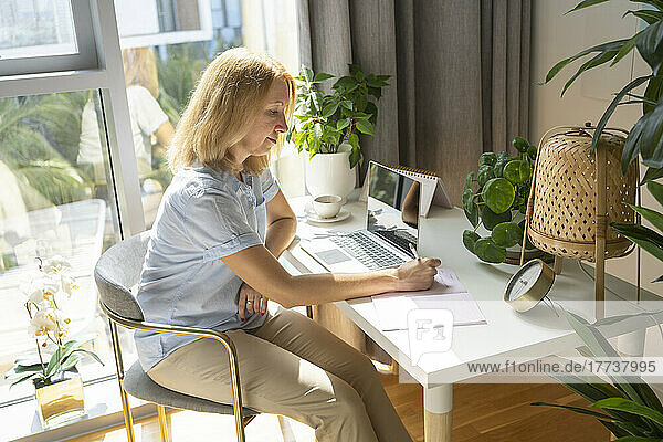Blond freelancer writing in dairy sitting on chair at desk in home office