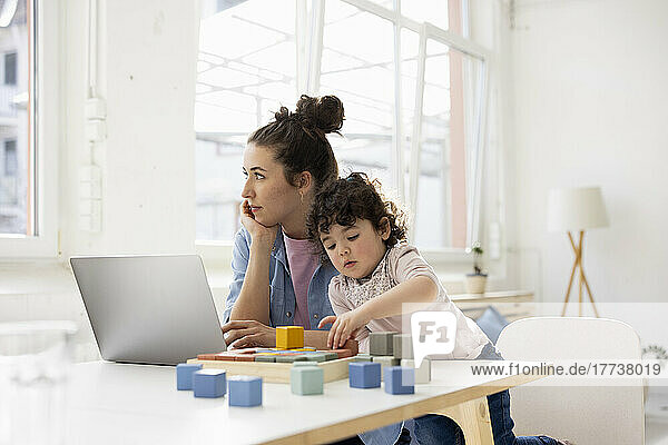 Mutter arbeitet von zu Hause aus mit Laptop  während Tochter mit Bauklötzen spielt