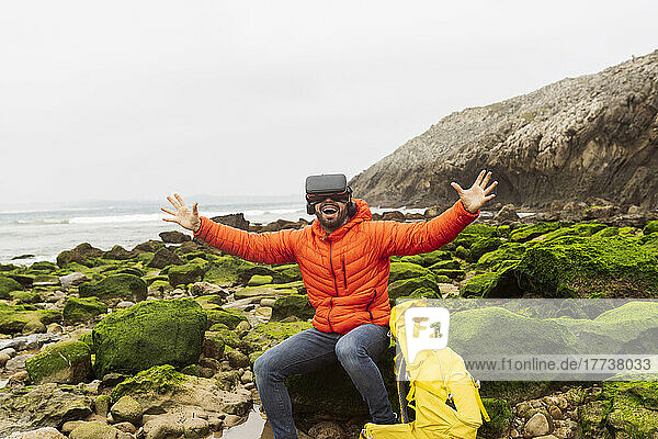 Glücklicher Mann mit Virtual-Reality-Simulator sitzt mit ausgestreckten Armen auf einem Felsen