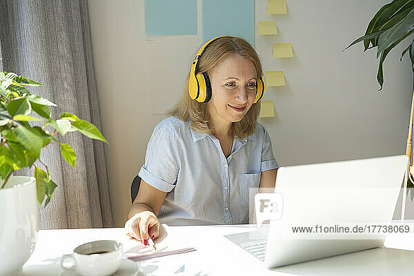 Smiling freelancer wearing wireless headphones using laptop at desk in home office