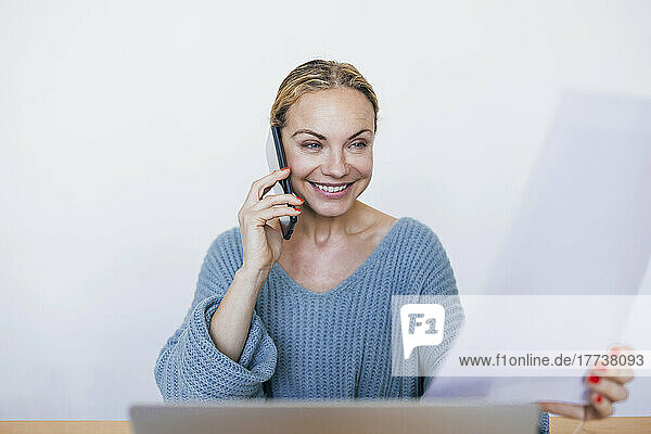 Smiling businesswoman holding document talking on mobile phone sitting with laptop at home