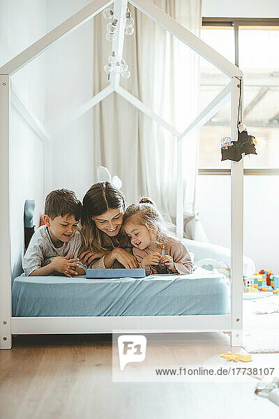 Smiling young woman with children looking at tablet PC lying on bed
