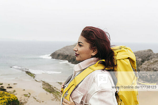 Lächelnde Frau mit roten Haaren  die einen Rucksack trägt und vor dem Meer steht
