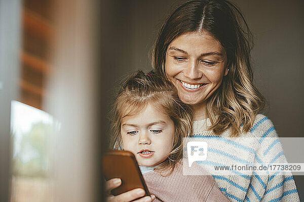 Lächelnde junge Frau zeigt ihrer Tochter durch das Fenster gesehen ihr Mobiltelefon