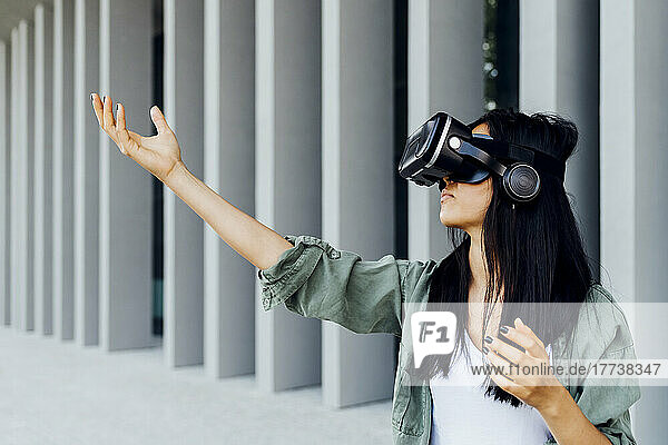 Young woman wearing virtual reality simulator gesturing in front of column