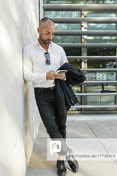 Man with jacket using smart phone leaning against wall