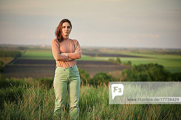 Confident young woman with arms crossed standing on field at sunset