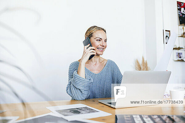 Happy businesswoman talking on mobile phone sitting with laptop at home