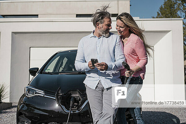 Smiling man showing mobile phone to woman holding electric plug on sunny day