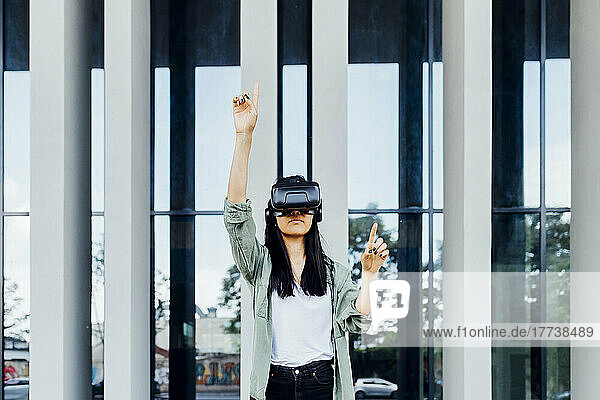 Young woman wearing virtual reality simulator standing with hand raised in front of column