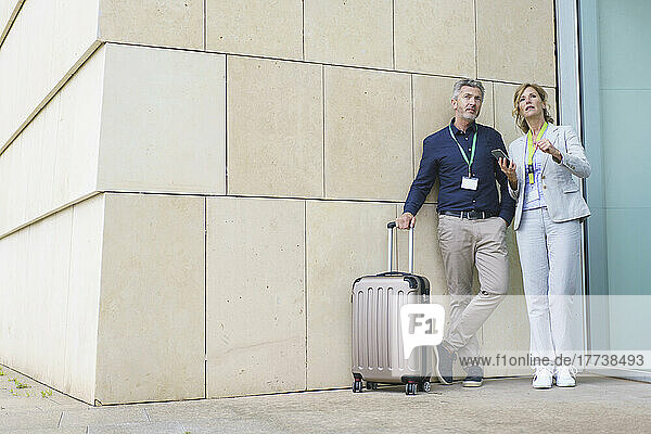 Businesswoman holding mobile phone discussing with businessman standing by suitcase in front of wall