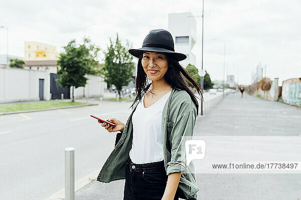 Smiling young woman wearing hat standing with smart phone on road