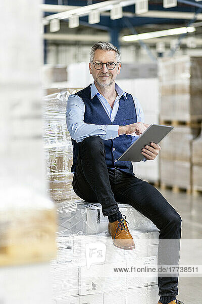Smiling manager with tablet PC sitting on boxes in warehouse