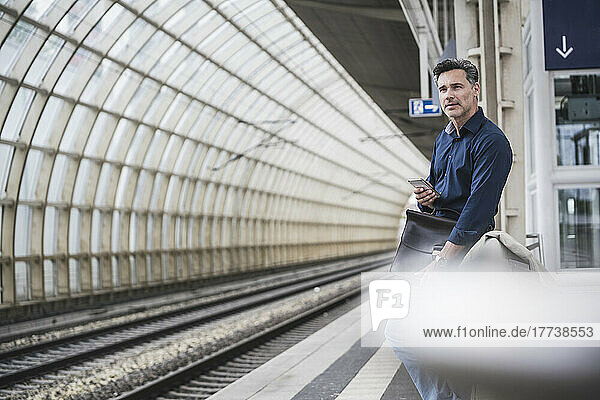 Businessman with smart phone and bag standing at railroad station